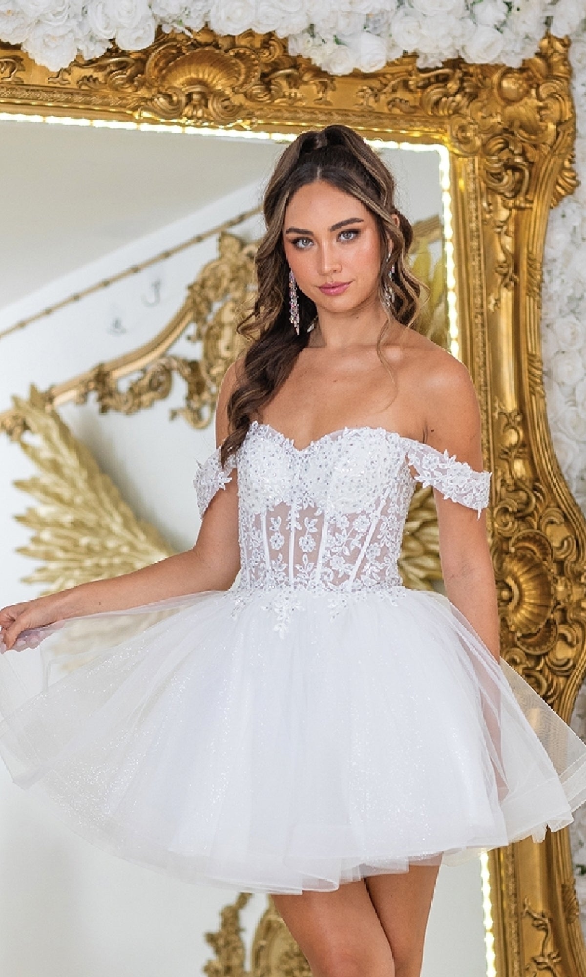 A woman in the Dancing Queen 3418 off-shoulder short prom dress with a fitted lace bodice and tulle skirt stands before a gold ornate mirror and white floral backdrop—an affordable choice for any special occasion.